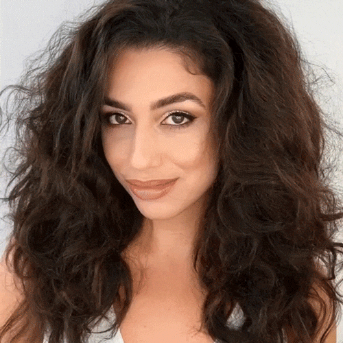 Smiling woman with long dark hair sectioned and clipped up, preparing to demonstrate how to use the beachwaver pro curling iron on camera
