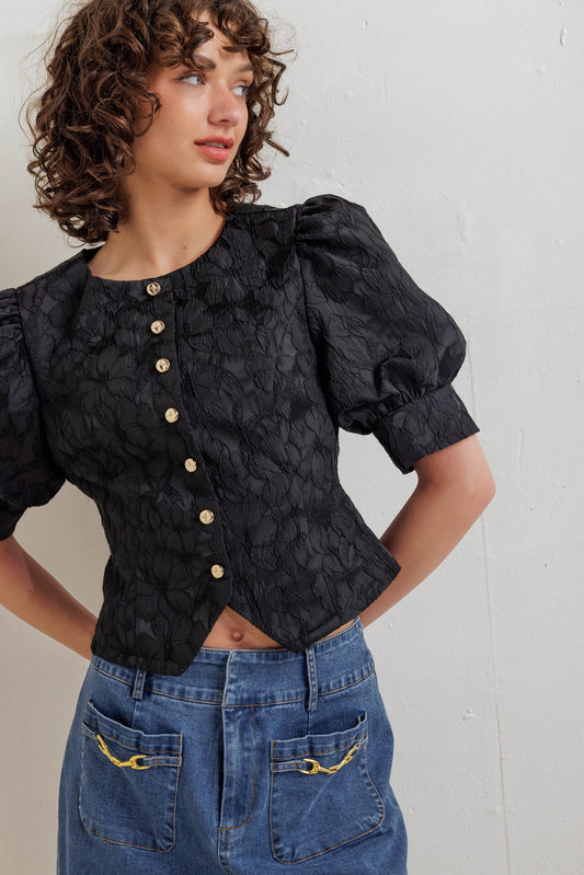 Woman wearing a black textured blouse with puff sleeves and blue jeans against a plain background