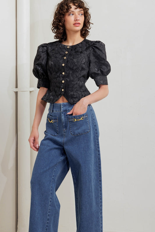 Woman wearing a black patterned blouse and blue jeans against a light gray background