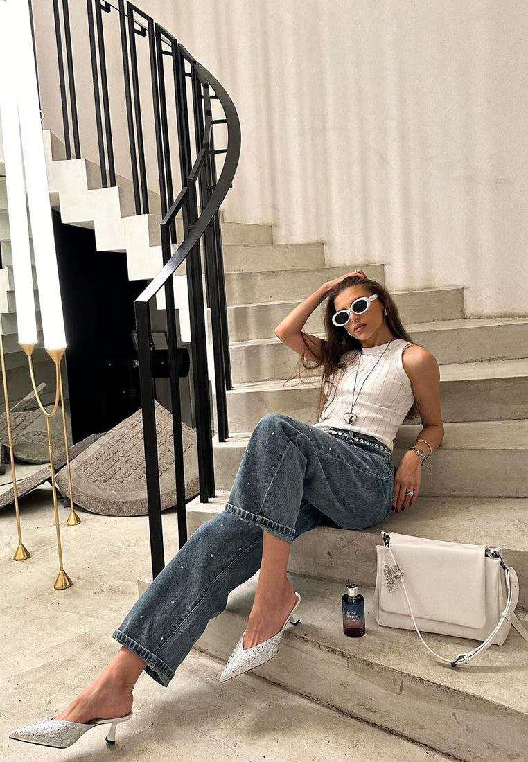 Woman sitting on a staircase wearing a white top, blue jeans, and silver high heels, with a white handbag.