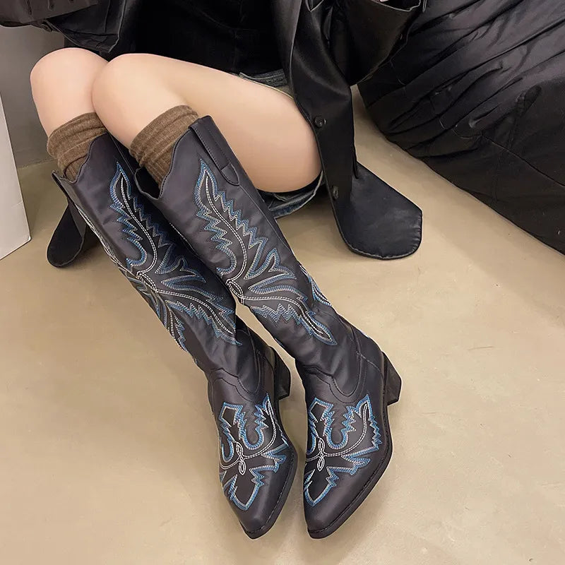 Person wearing black knee-high boots with blue embroidery on a beige floor.